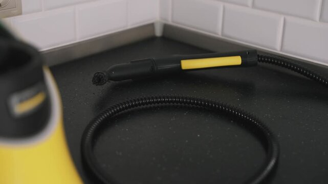 Close up shot of a yellow and black steam cleaner nozzle resting on a black speckled kitchen countertop with white subway tile backsplash visible in the background