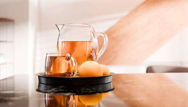 A glass pitcher and a cup of tea with oranges on a tray on a reflective table.