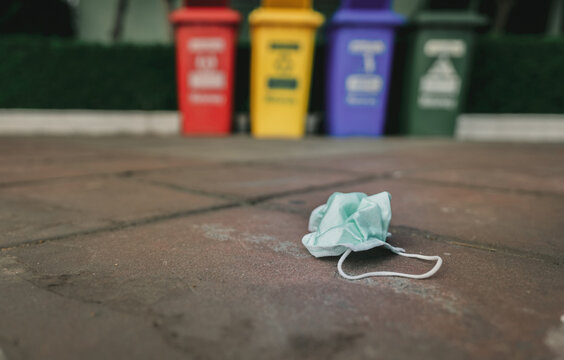 Used medical face mask discard on pavement floor on blurred recycle bin or trash. Medical waste disposal with unhygienic. Disposable earloop face mask waste garbage on floor. Green surgical face mask.