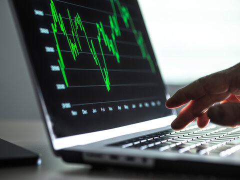 Hand pointing at laptop screen displaying green financial graph in a dimly lit room.