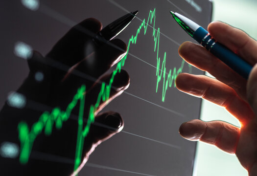 Hands pointing at a stock market graph on a screen with green line chart. 