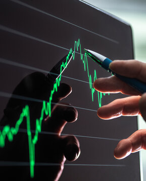 Hand holding a pen pointing at a green financial chart on a screen.