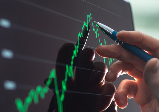 Hand holding a pen pointing at a financial chart on a computer screen.