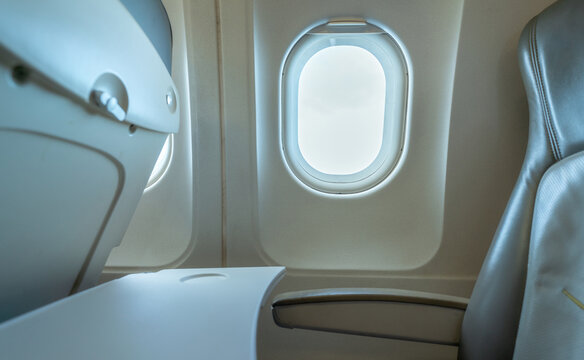 Plane window with white sunlight. Empty plastic airplane tray table at seat back. Economy class airplane window. Inside of commercial airline. Seat with armchair. Leather seat of economy class plane.
