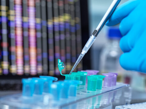 Gloved hand using pipette with colorful test tubes in a lab setting with data screen behind.