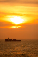 Fototapeta premium A cargo ship sails under a vibrant orange sunset on a calm ocean horizon. Vung Tau, Vietnam
