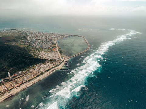 Aerial view of coastal town with waves crashing onto rocky shoreline and a protective sea wall. Ly Son island, Vietnam