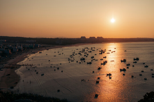 Sunset over a serene bay with numerous boats, casting reflections on the water surface.