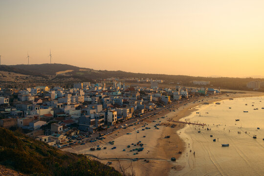 Coastal town at sunset with wind turbines and people on a sandy beach.