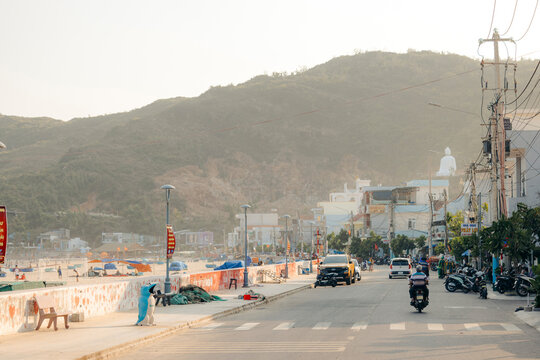 Scenic coastal street with mountains and people, vehicles, and shops in warm evening light.