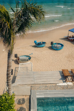 A tranquil beach scene with boats on the sand, palm tree, and adjacent wooden deck and pool.