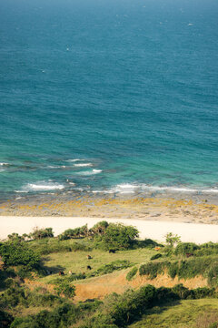 Coastal landscape with vibrant blue sea, sandy beach, and green hills in sunlight.