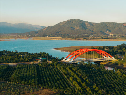 Red arch bridge spanning over a river with lush green fields and mountains in the background.