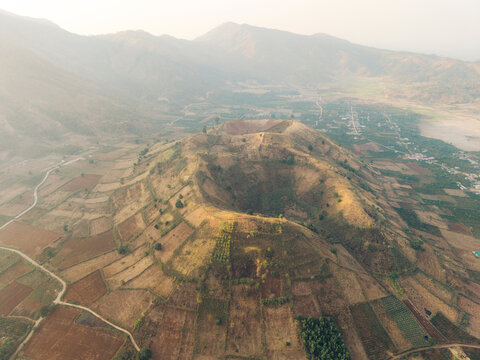 Aerial view of a crater amidst patchwork farmland with a hazy mountainous backdrop.