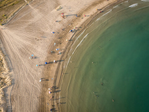 Aerial view of a sandy beach with scattered people and umbrellas along clear turquoise water.