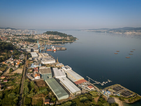 Aerial view of an industrial port city with lush greenery and calm sea on a clear day.
