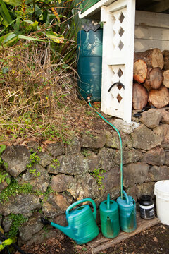 Three green watering cans on the ground by a stone wall with logs and a garden hose nearby.