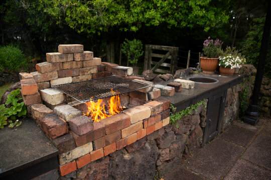 Brick barbecue grill with flames in a garden setting, surrounded by lush greenery.