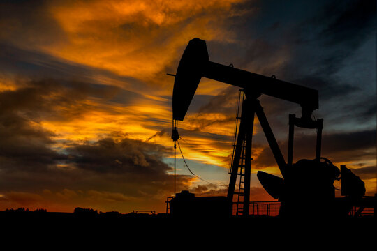 Silhouetted oil pump jack against a dramatic sunset sky with vibrant orange clouds. Canada