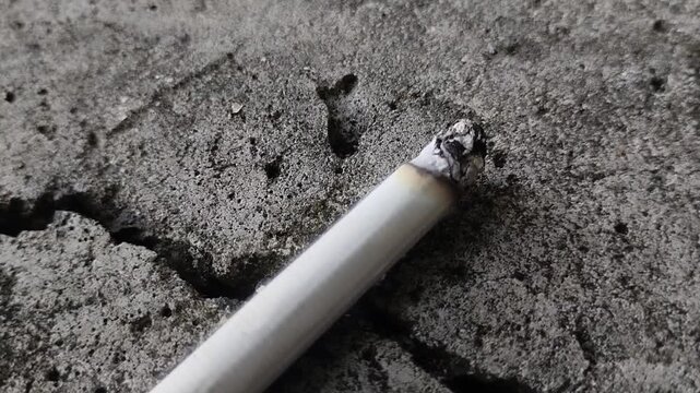 A cigarette butt glows with a thin layer of smoke on a cracked concrete floor. This concept reflects the dangers of smoking, addiction, and the negative impacts of environmental pollution