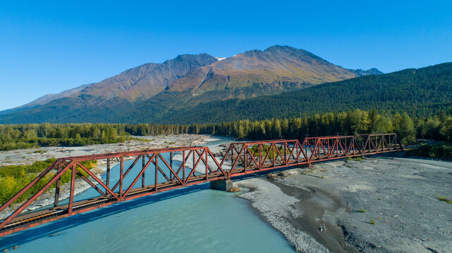 Rusty red bridge spans a wide river with majestic mountains and forests in the background. Moose Pass, Alaska, USA