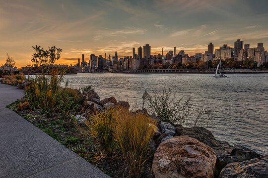 Sunset city skyline view from a riverside path with rocks and plants; a sailboat on the river. New York, USA