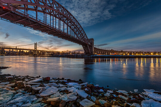 Majestic arched bridge spans a calm river at sunset, with city skyline and reflections below. New York, USA