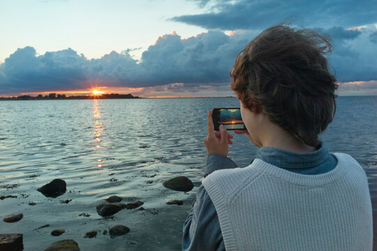 A person photographs a vibrant sunset over a calm sea with a smartphone on a rocky shore.
