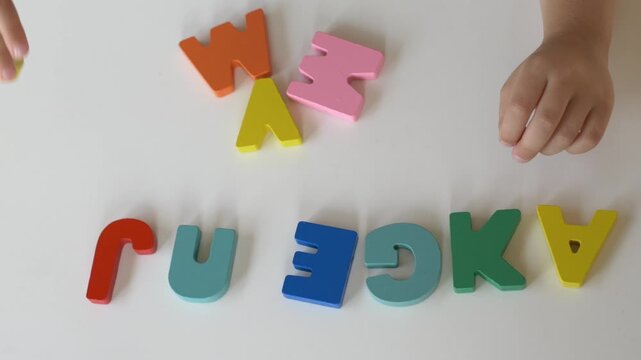 Child arranges colorful alphabet characters on white table while learning letter shapes and language fundamentals. Educational game develops literacy and recognition skills.