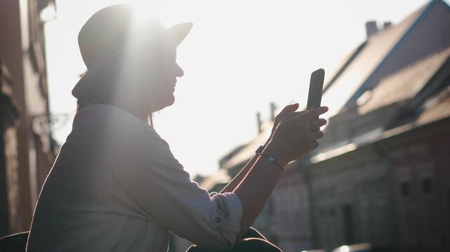 Female traveler in a hat looking at her smartphone while sitting on the steps of a historic building in a European city during a beautiful sunset.