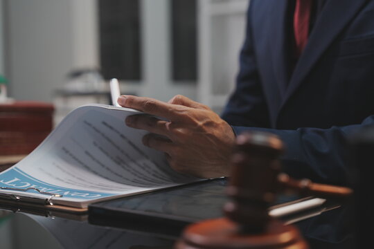 A dramatic, selective-focus close-up shot of a wooden judge's gavel being struck down onto its sound block. In the blurred background, a lawyer in a dark suit sits at a polished desk.  