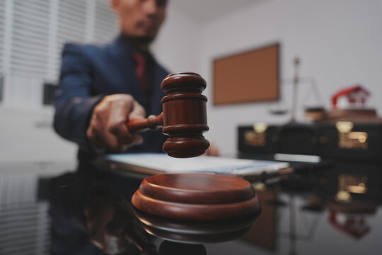 A dramatic, selective-focus close-up shot of a wooden judge's gavel being struck down onto its sound block. In the blurred background, a lawyer in a dark suit sits at a polished desk.  
