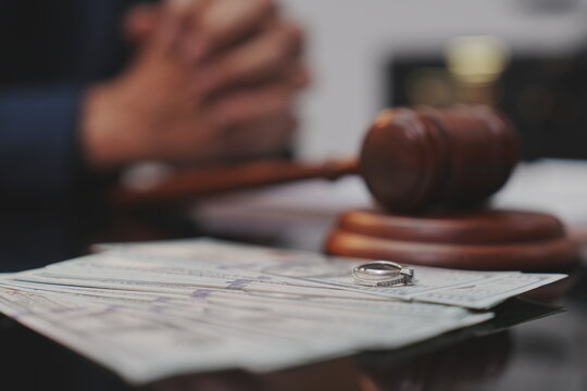A dramatic, selective-focus close-up shot of a wooden judge's gavel being struck down onto its sound block. In the blurred background, a lawyer in a dark suit sits at a polished desk.  