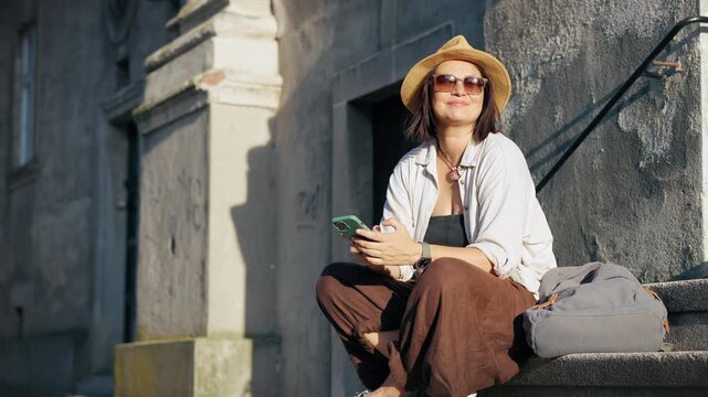 Female traveler in a hat looking at her smartphone while sitting on the steps of a historic building in a European city during a beautiful sunset.
