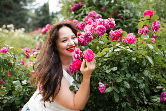 Happy young adult woman 30-35 year old with red lipstick make up and long hair in blooming rose bushes with flowers in garden outdoors close up. Summer vacation season. Healthy lifestyle.