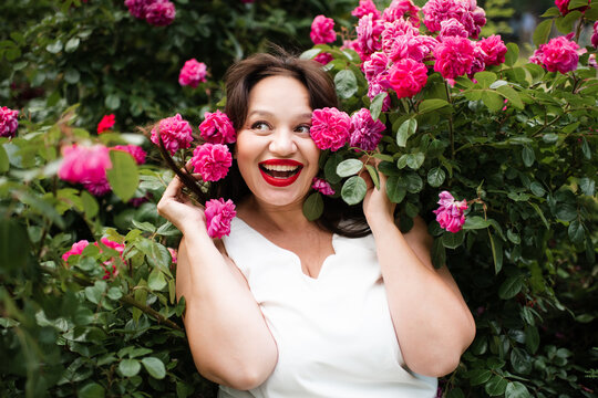Cheerful laughing adult woman 30-35 year old with red lipstick make up and long hair in blooming rose bushes with flowers in garden outdoors close up. Summer vacation season. Positive emotions.