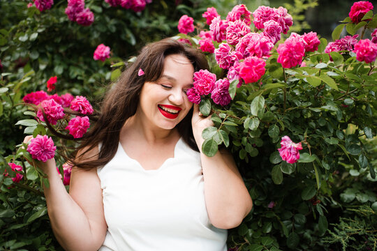 Smiling young adult woman 30-35 year old with red lipstick make up and long hair posing in blooming rose flowers in garden outdoors close up. Summer vacation season. Healthy lifestyle.