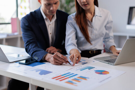Two asian confident business people point to graphs and charts to analyze market data, balance sheet, account, net profit to plan new sales strategies to increase production capacity Financial and Acc