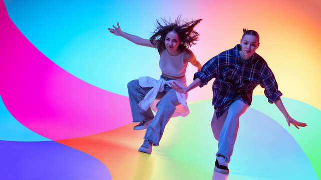 Two female dancers moving in colorful neon studio with abstract background. Concept of dance crew, youth lifestyle, music promotion, street fashion, and creative advertising.