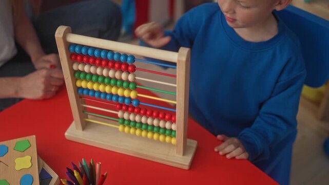 Boy pushes beads on wooden abacus with hand at red table inside building while adult hand guides; concentration learning.