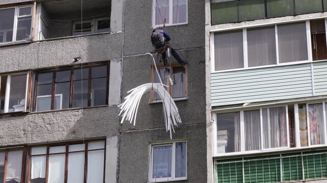 Worker suspended at height performs facade maintenance, applying insulation to seal gaps between concrete panels.