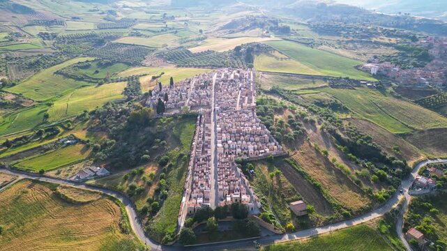 Aerial view of a historic cemetery with rows of tombs surrounded by rolling green hills and agricultural fields in Santa Caterina Villarmosa, Sicily, Italy.
