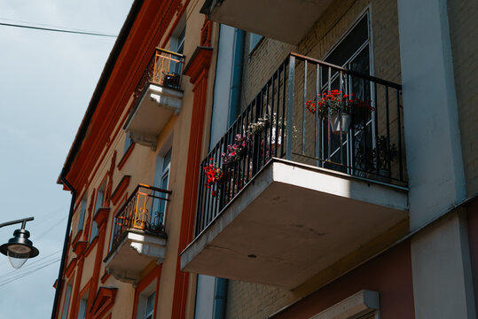A balcony is adorned with various flowering plants, adding color to a building's exterior. Sunlight shines on the vibrant flowers, creating a bright atmosphere in the urban setting