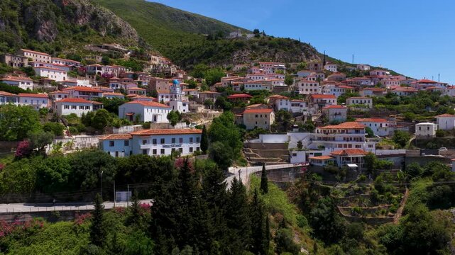 Aerial view of the traditional white houses and red-tiled roofs of Dhermi village nestled on a lush green hillside under a clear blue sky in Dhermi, Vlore County, Albania.