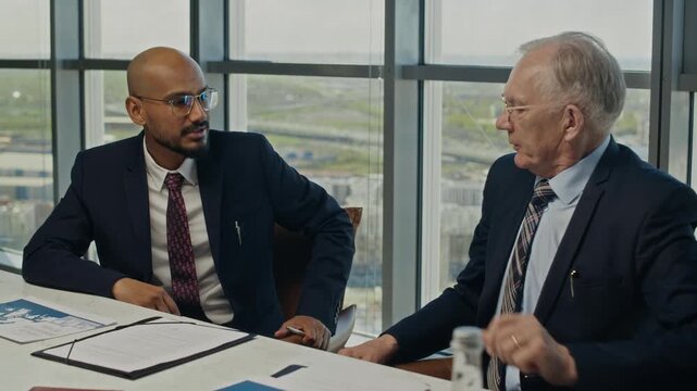 Tilt shot of Indian businessman and senior Caucasian businessman discussing project, standing up to shake hands and leaning over documents. Ideal for corporate partnership and energy sector videos