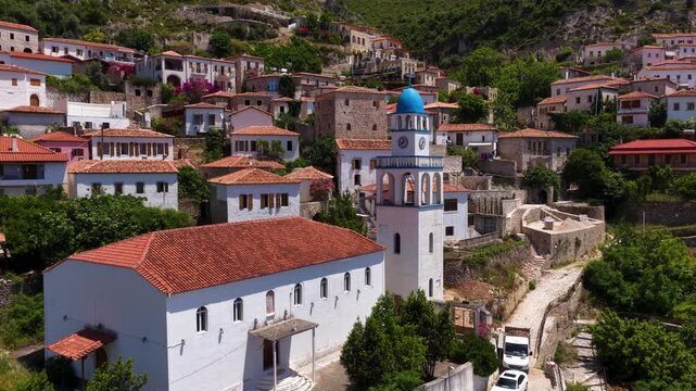 Aerial view of a white church with a blue dome and bell tower surrounded by traditional stone houses and lush green hills in Dhermi, Vlore County, Albania.