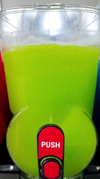 Vertical video social media - Closeup of a generic unbranded commercial icy drinks machine, with bright green slush textured contents being agitated, ready to deliver a semi frozen, sweet beverage.
