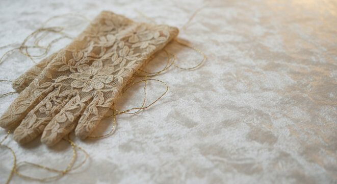 Delicate beige lace gloves with floral patterns and gold thread detail resting on a textured white fabric surface, elegant vintage accessory close-up