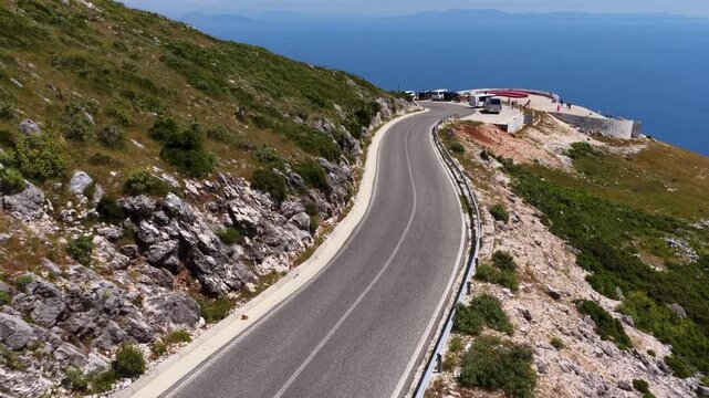 Aerial view of the winding Llogara Pass mountain road curving along steep rocky slopes with people at a viewpoint overlooking the Ionian Sea in Llogara, Vlore County, Albania.