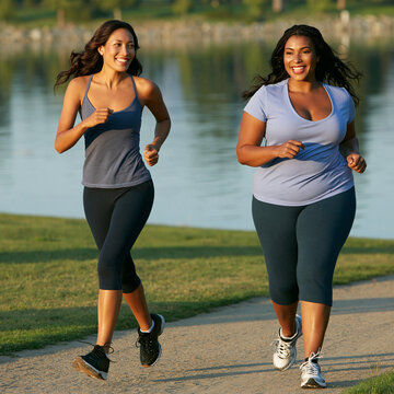 Two women of different body types running outdoors in a park next to a lake. They are both smiling and appear to be enjoying their workout